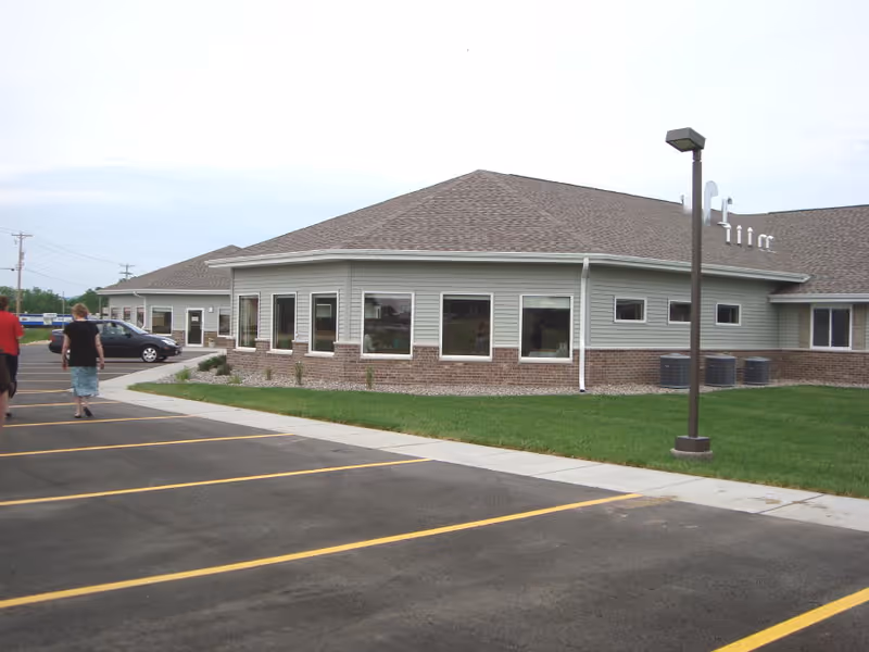 Exterior view of a single-story building with beige siding and brick accents, multiple windows, a parking lot with yellow lines, a sidewalk, a street lamp, and three people walking near the building.
