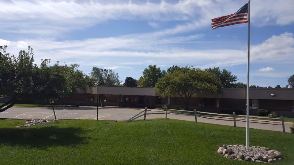 Exterior view of Medilodge of Westwood facility with a green lawn in the foreground, an American flag on a flagpole to the right, trees, and a single-story building under a partly cloudy sky.