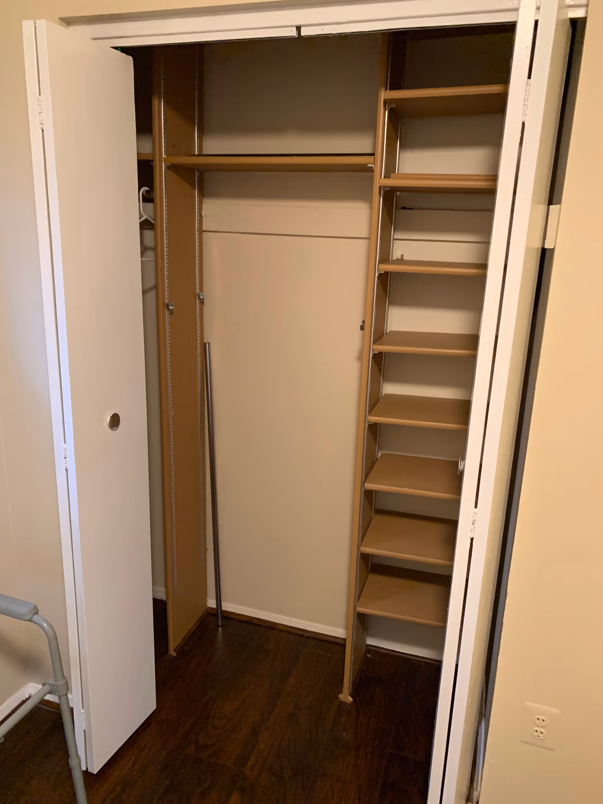 An empty closet with white bi-fold doors partially open, featuring wooden shelves on the right side and a hanging rod on the left. The closet has a dark wooden floor and beige walls.