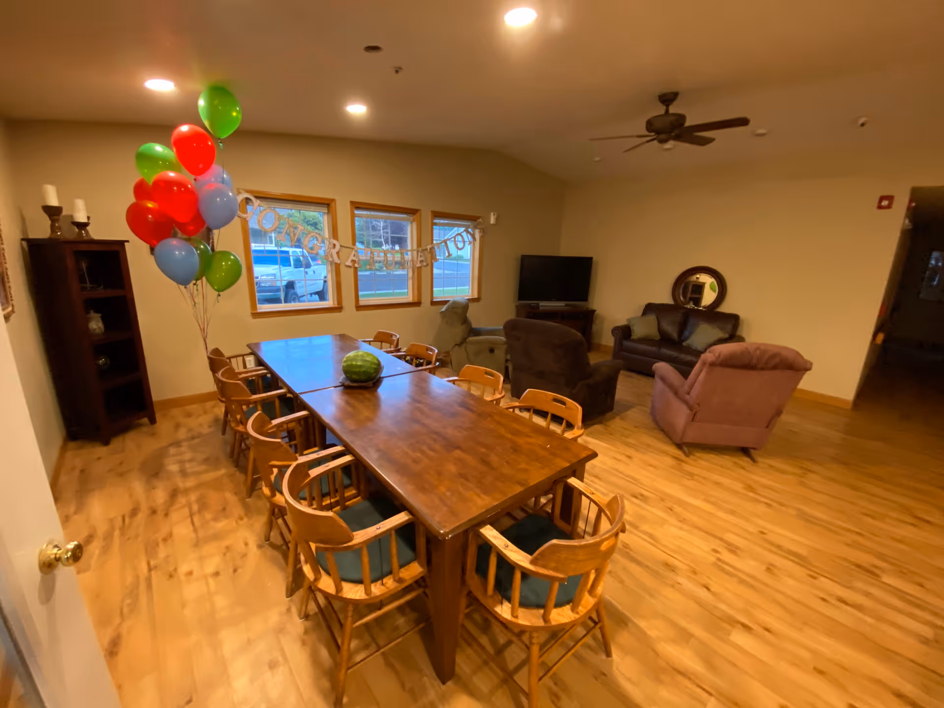 Communal dining and lounge area with a long wooden table surrounded by chairs, balloons and a 'Congratulations' banner, plus sofas and a TV.
