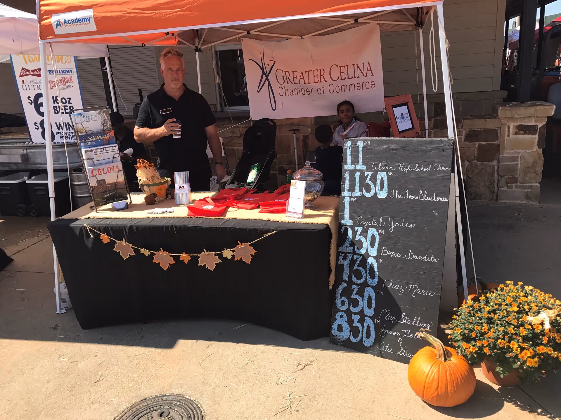 Two people at an outdoor booth under an orange canopy with a banner and a pumpkin.