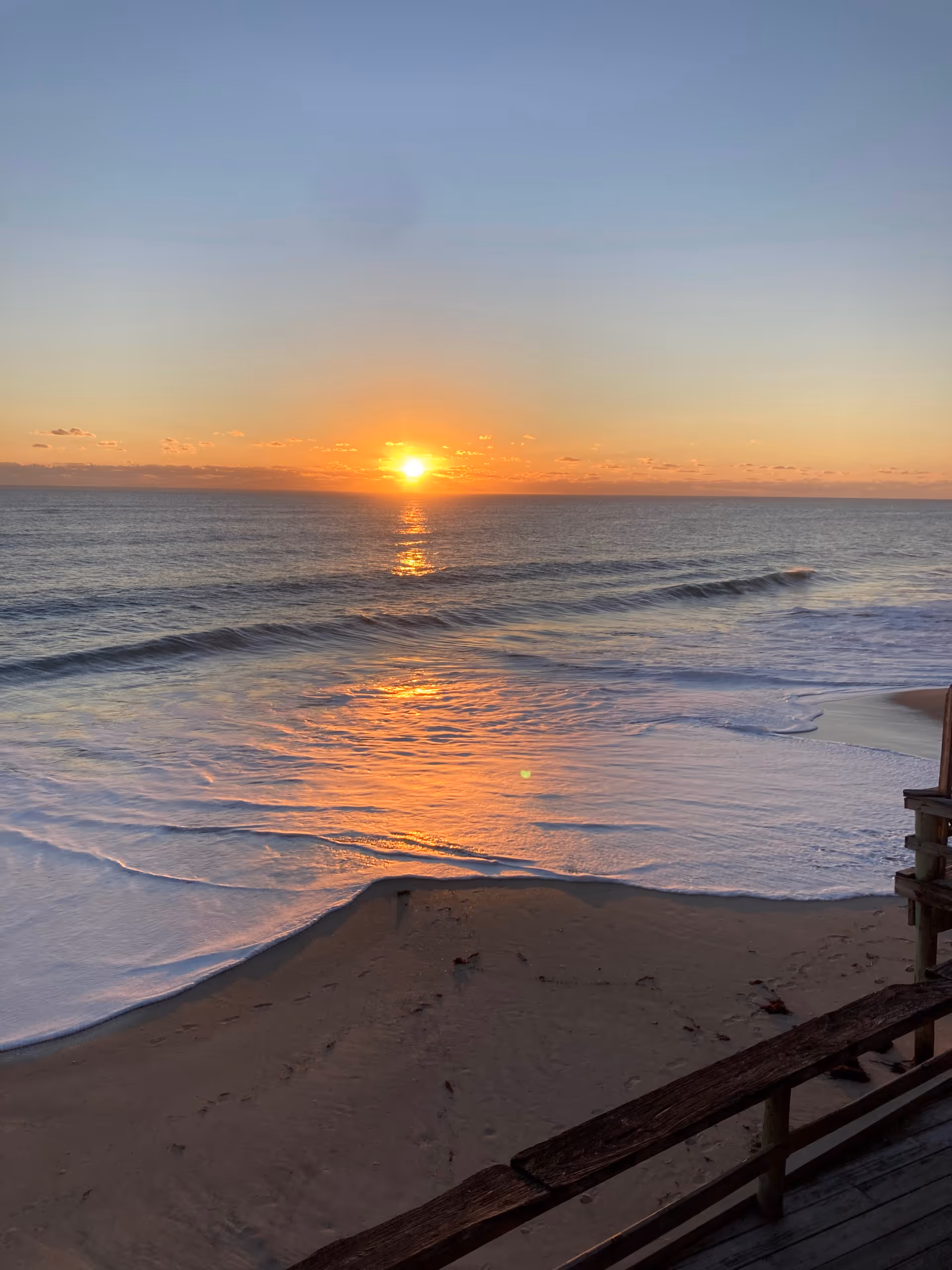Sunset over the ocean with waves, sandy beach, and a wooden boardwalk railing.