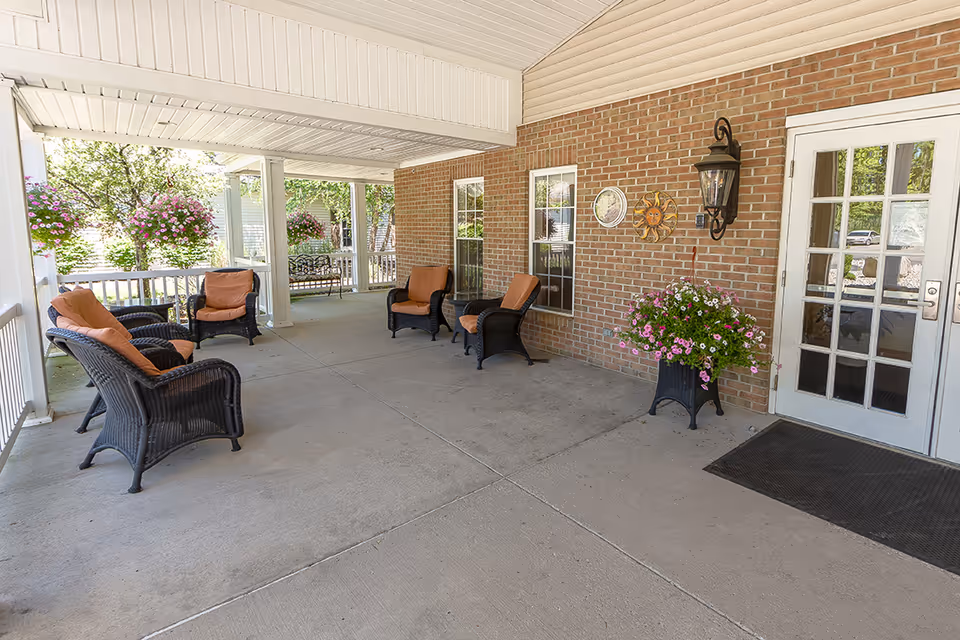 Covered front porch of an assisted living facility with wicker chairs, potted flowers, and a glass door entrance.