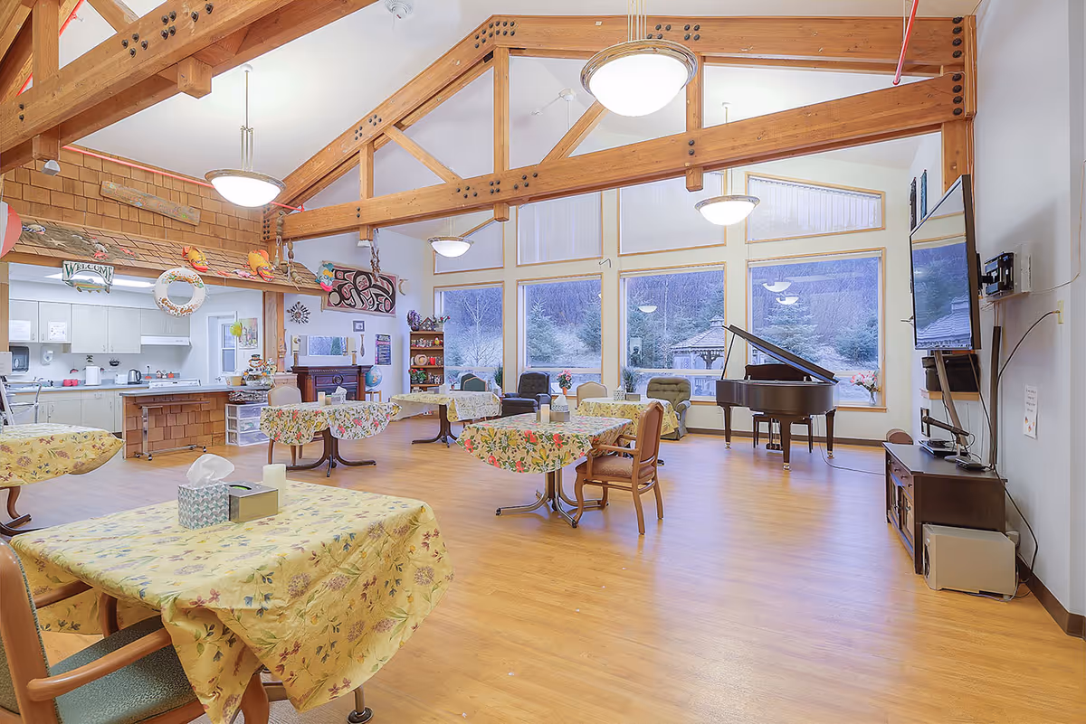 A spacious, well-lit common area with wooden beams on the ceiling, several tables covered with floral tablecloths, chairs, a grand piano near large windows showing an outdoor view, and a kitchen area in the background.