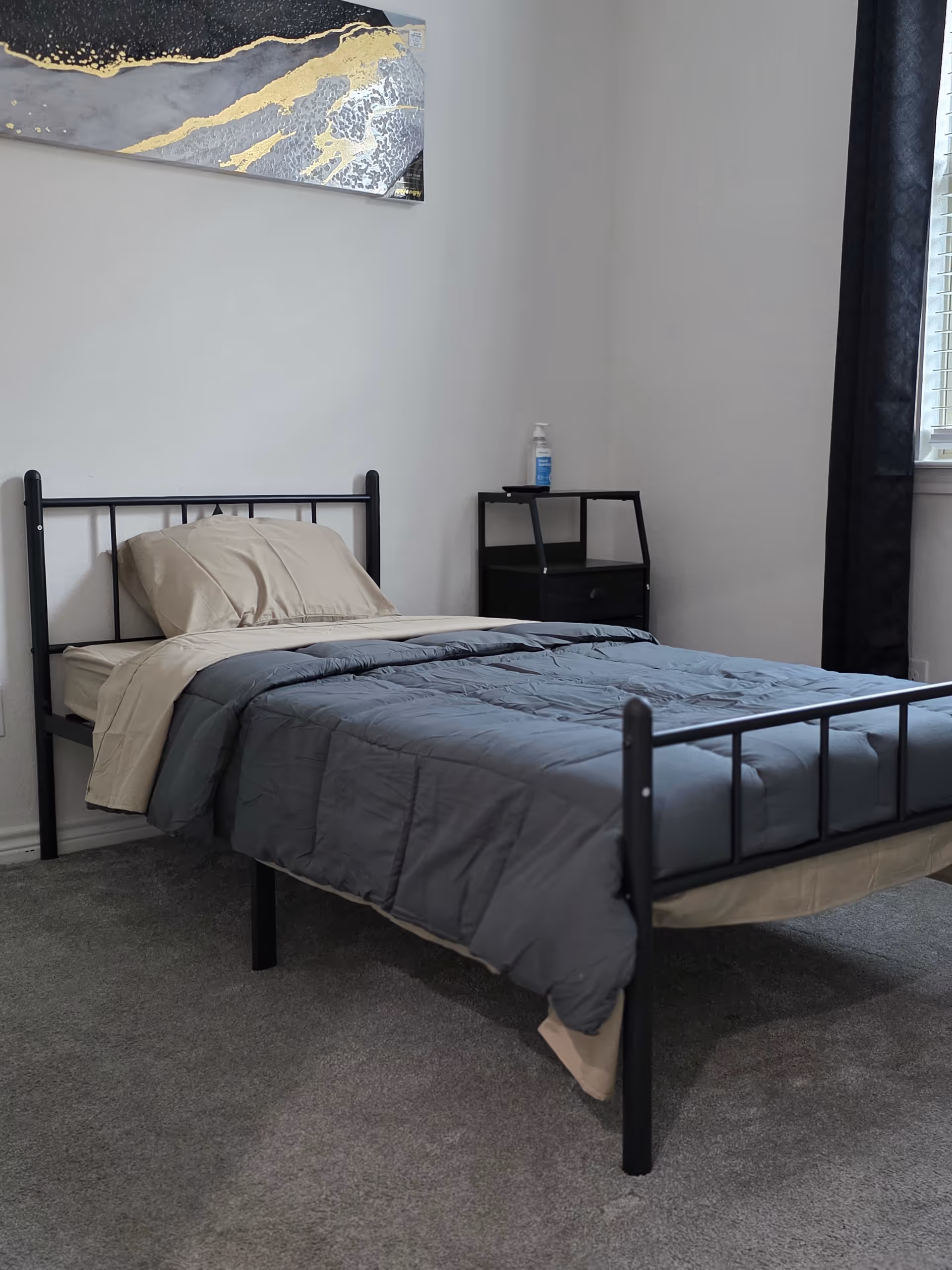 A simple bedroom with a metal bed frame, gray comforter, beige sheets, a nightstand, and window.