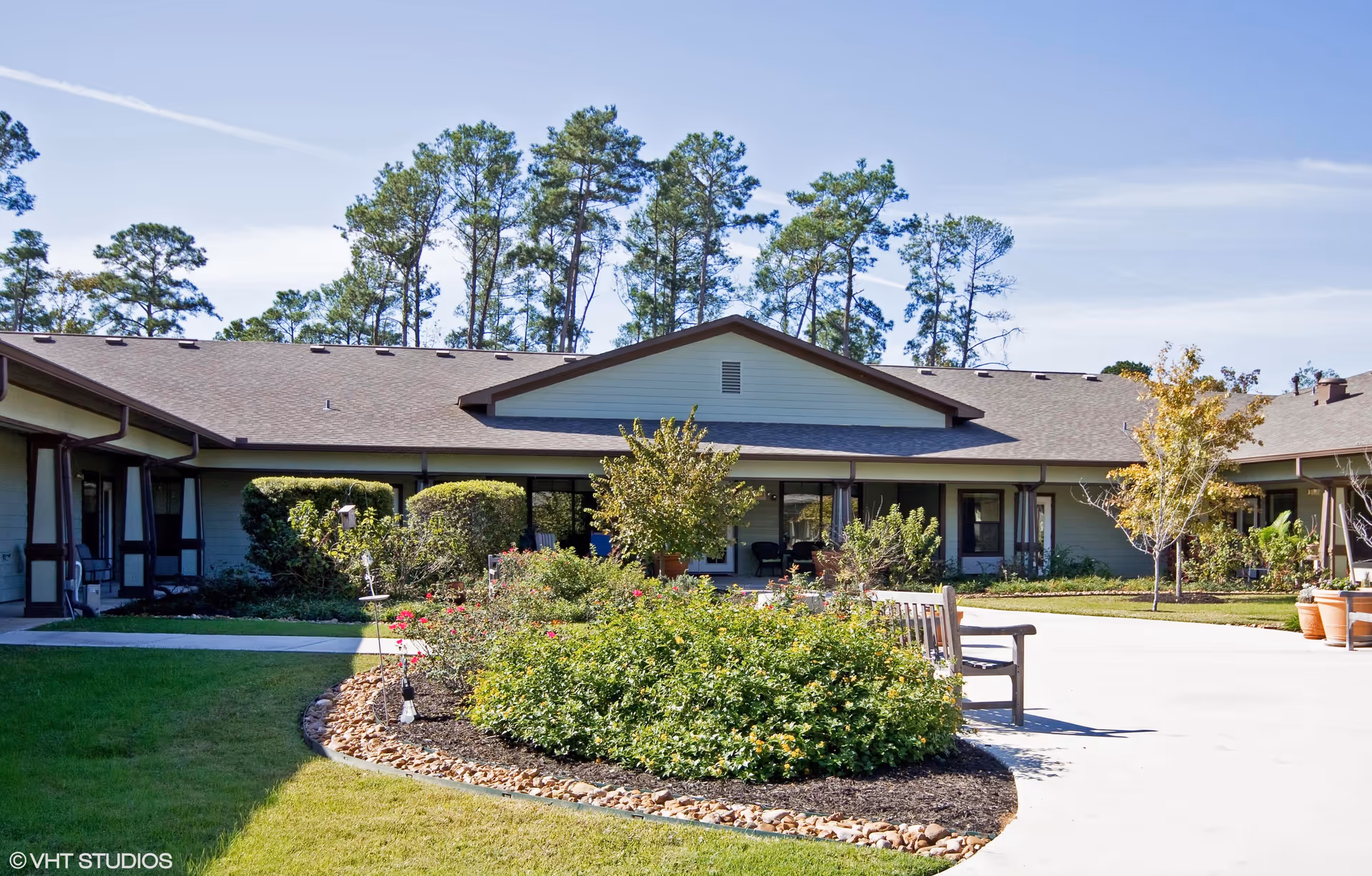 Exterior view of a single-story assisted living facility building with a landscaped garden in front, including bushes, flowers, and a wooden bench. Tall pine trees are visible in the background under a clear blue sky.