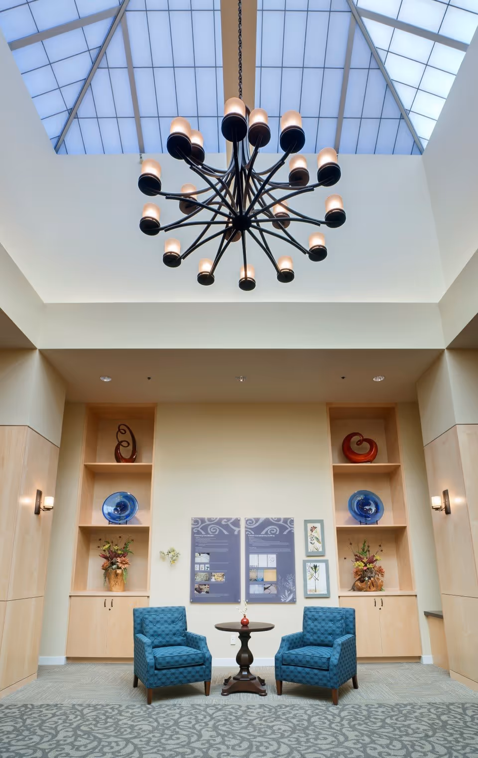 A bright interior seating area with two blue patterned armchairs and a small round wooden table between them. Behind the chairs are built-in wooden shelves with decorative items including vases and plates. Above, a large modern chandelier hangs from a high ceiling with a skylight.