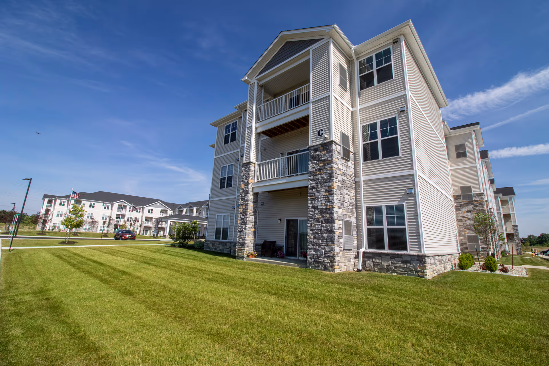 Front exterior of a multi-story senior living apartment building with balconies and a large lawn under a blue sky.