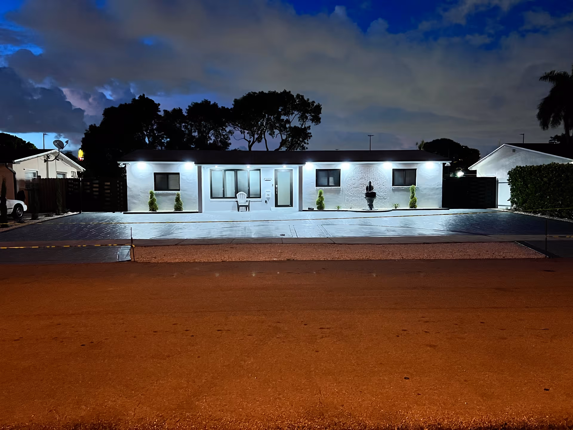 Front view of a single-story white building at night with exterior lights, a paved driveway, and a small fountain.