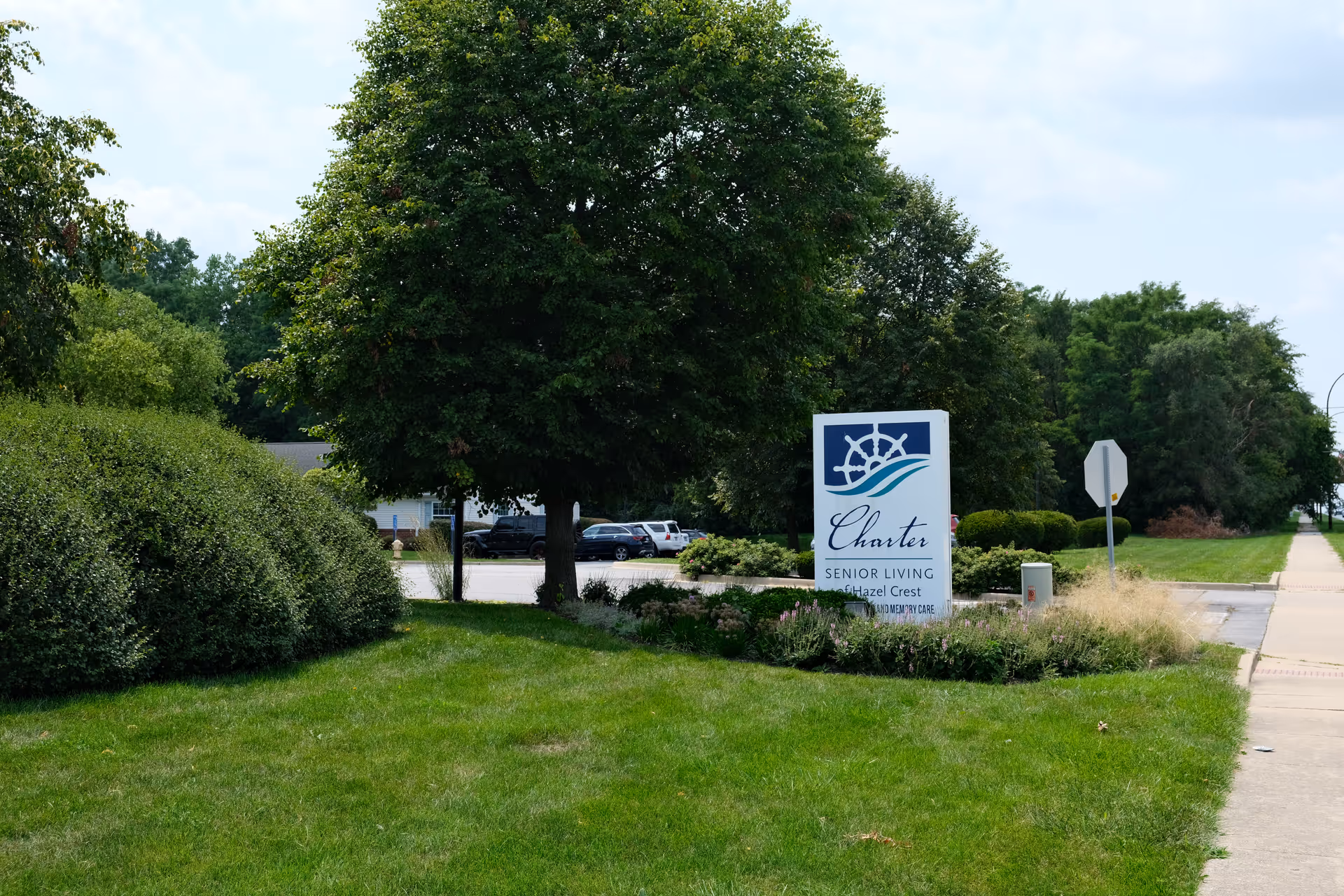 Front lawn and entry sign for Charter Senior Living of Hazel Crest next to trees, shrubs, and a sidewalk.