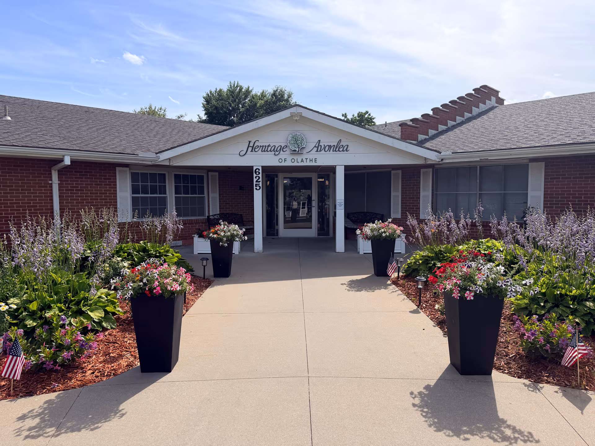 Front entrance of Heritage Avonlea of Olathe, surrounded by blooming planters and shrubs.