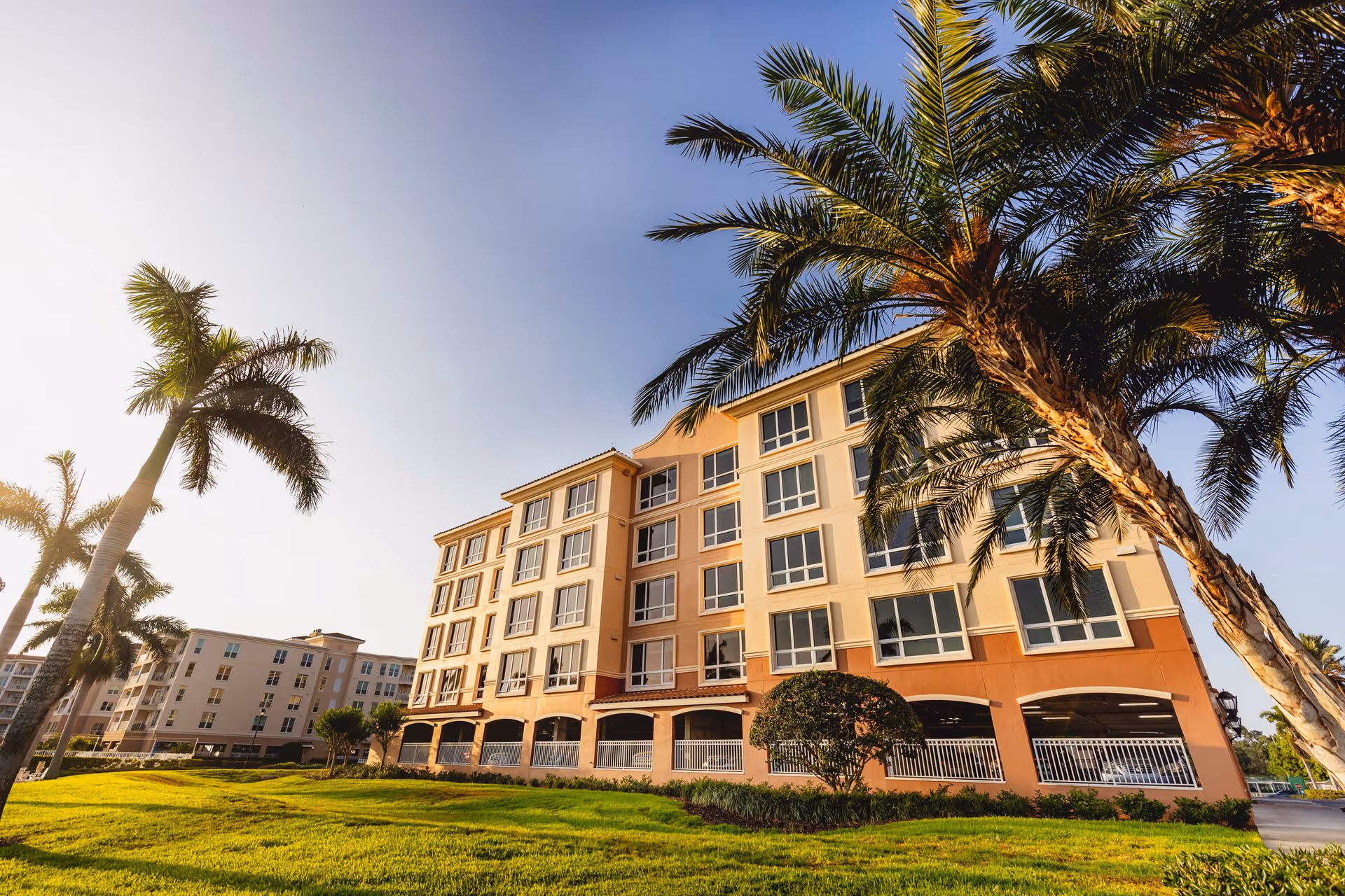 Exterior view of a multi-story senior living facility building with large windows, surrounded by palm trees and green grass under a clear sky.