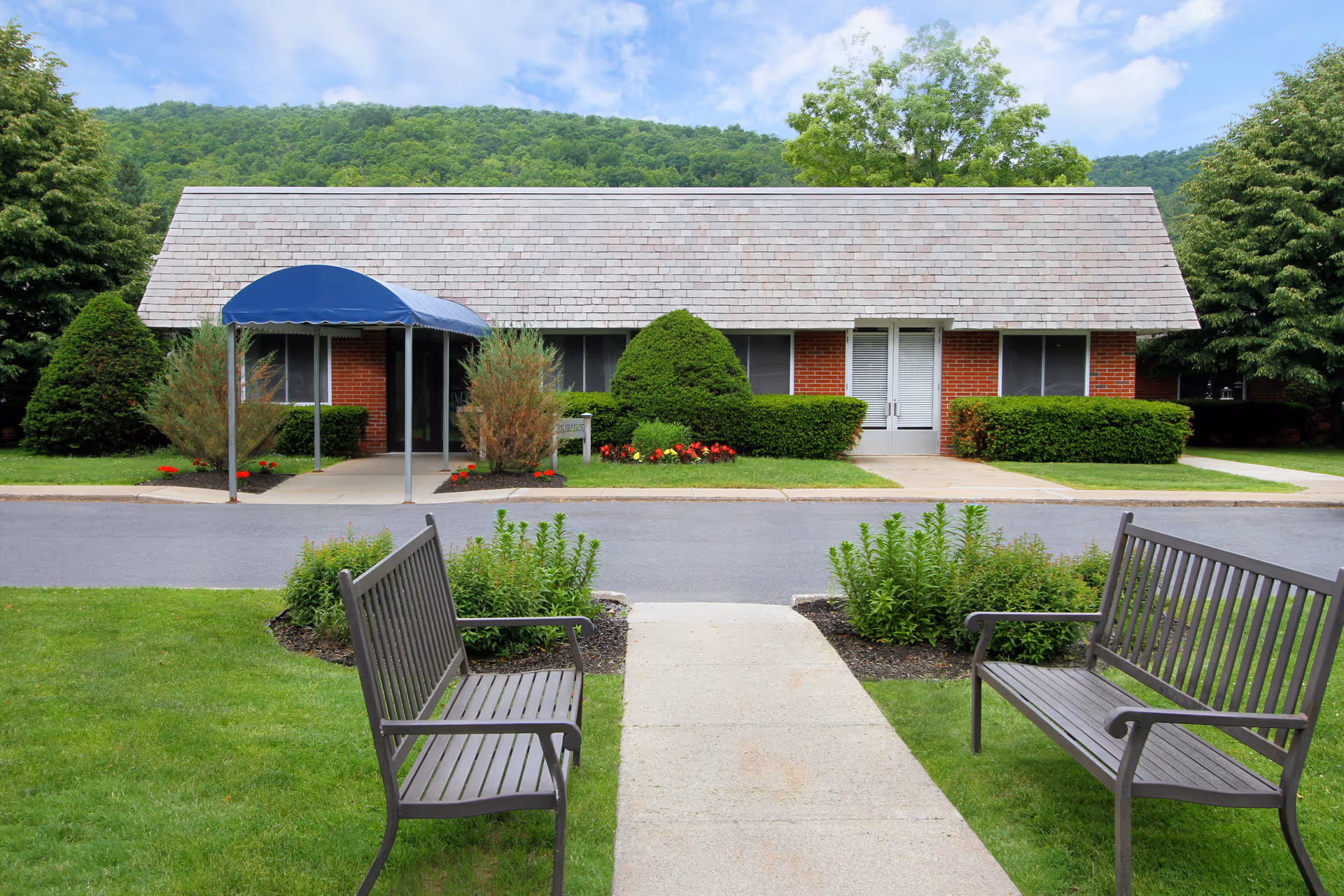 Exterior view of a brick senior facility with a blue canopy, benches, and manicured landscaping.