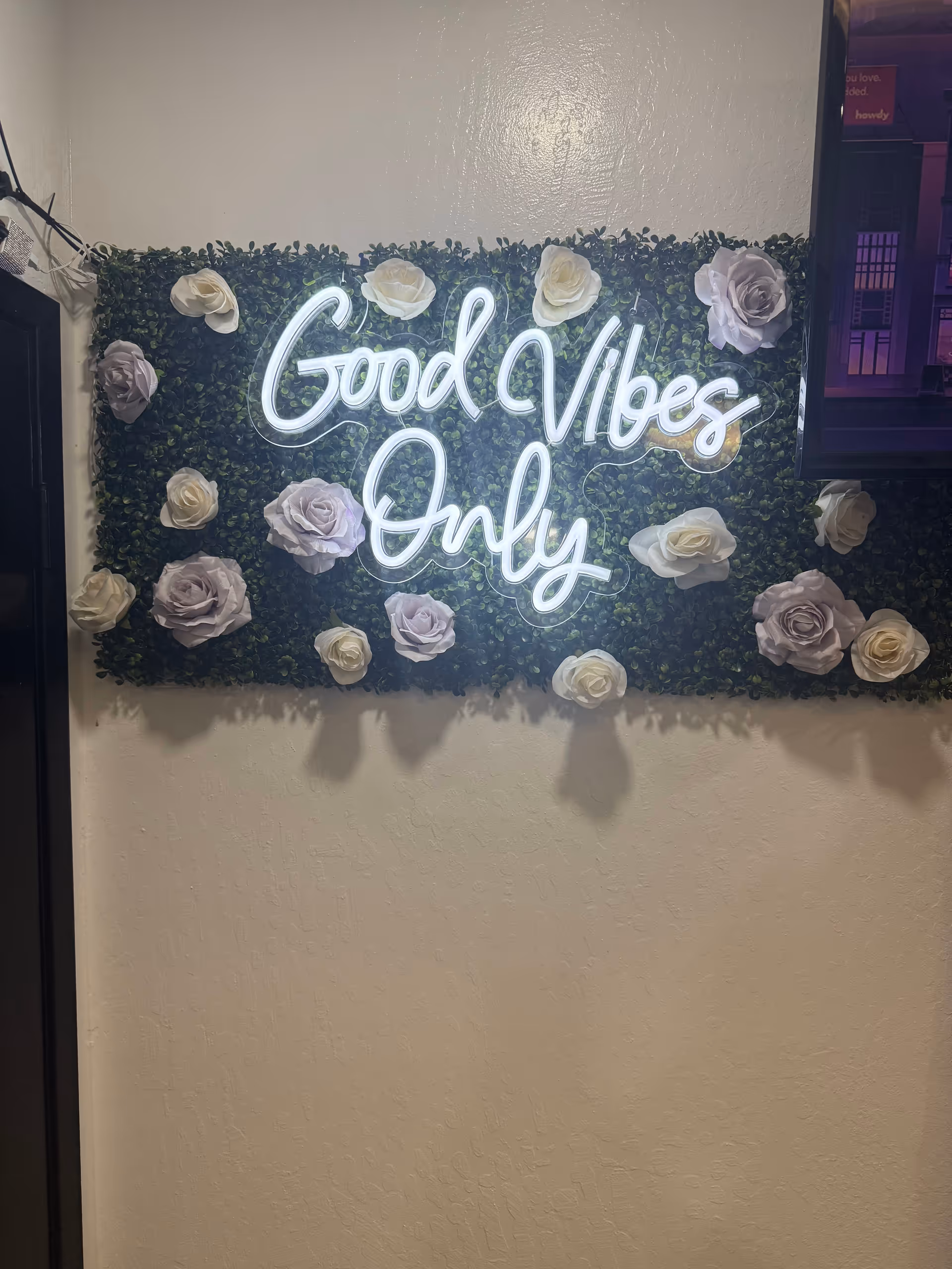 Neon sign reading Good Vibes Only on a faux greenery wall with roses.