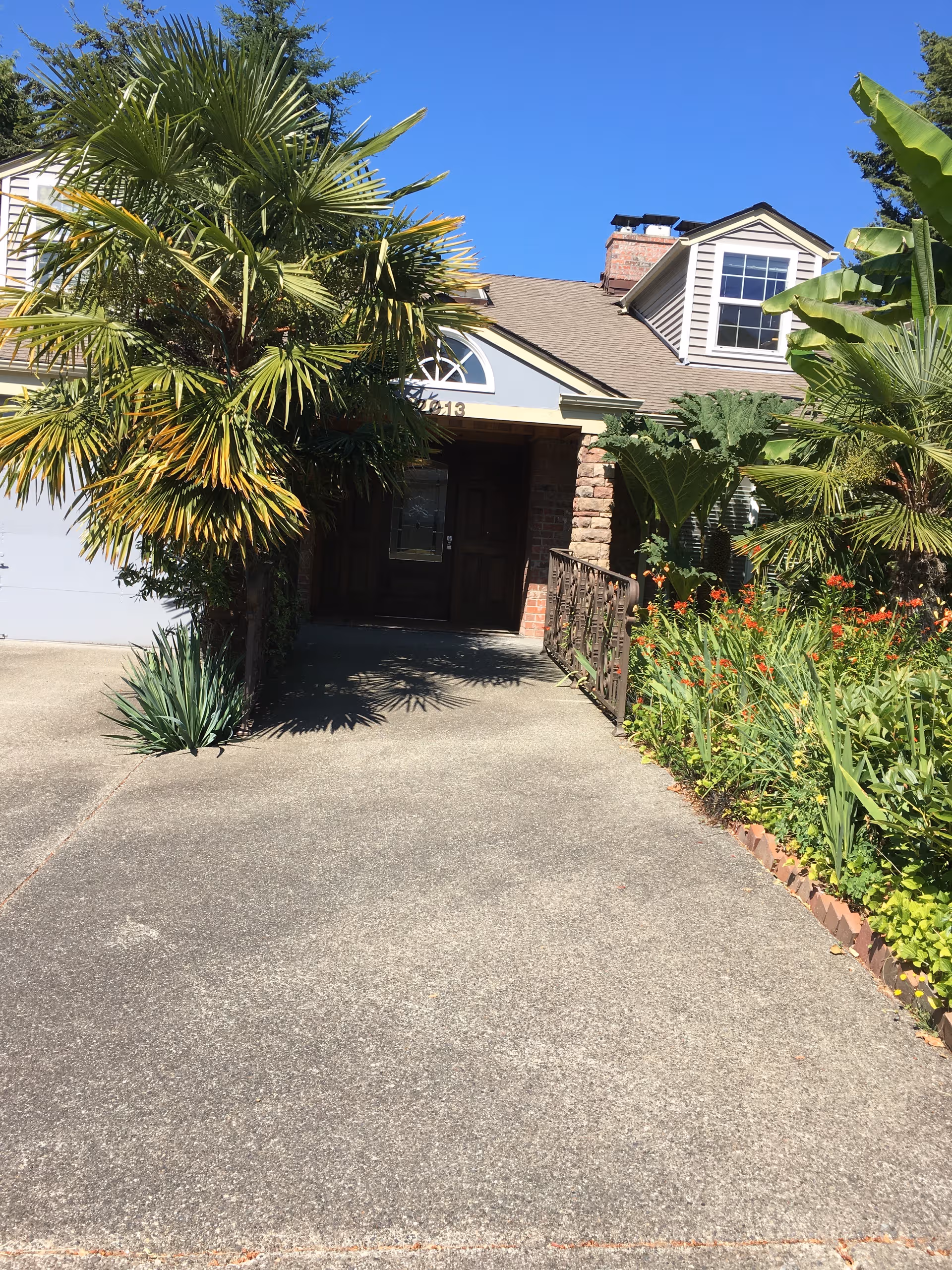 Sunny front entrance of a house-style senior home, surrounded by palm trees and lush landscaping.