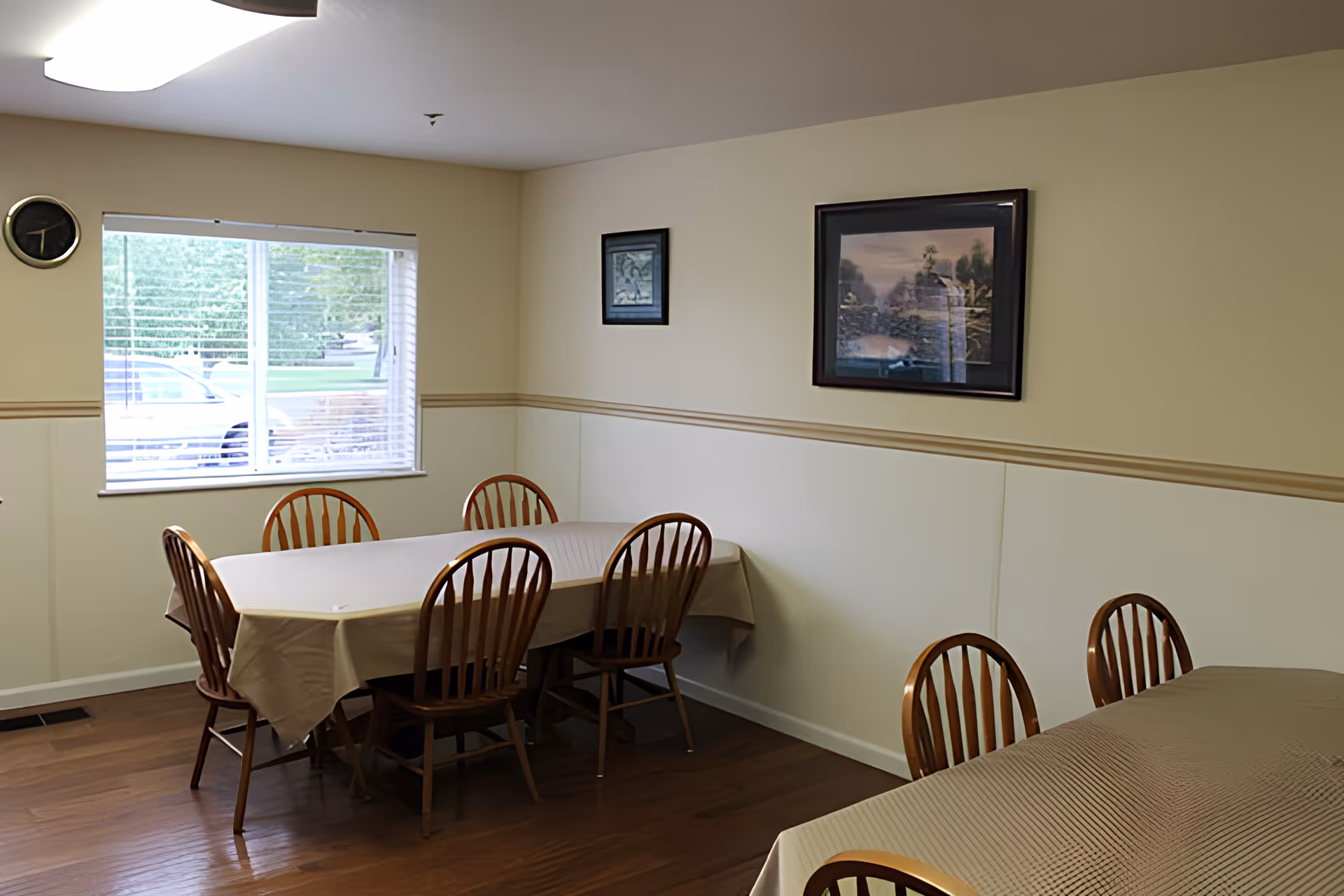 A small dining room with two tables draped in tablecloths surrounded by wooden chairs, a window letting in daylight, and framed artwork on the walls.