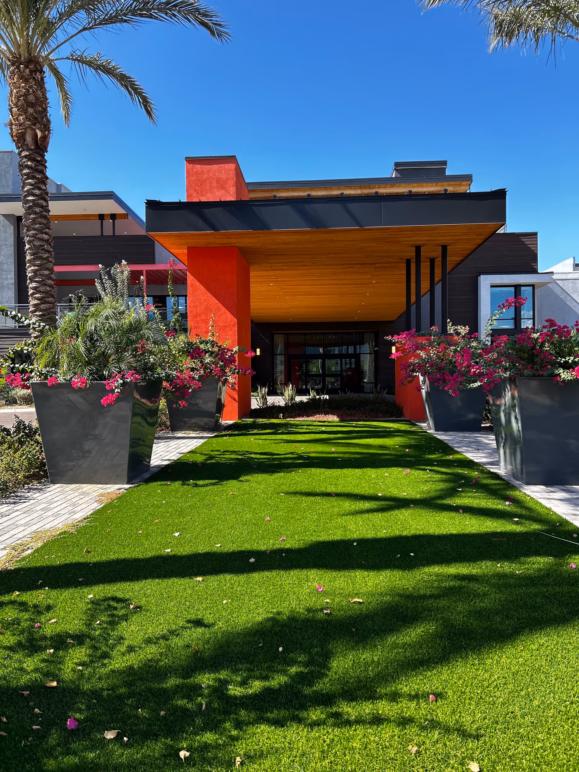 Modern facility entrance with red columns, wooden overhang, palm trees, and manicured lawn.