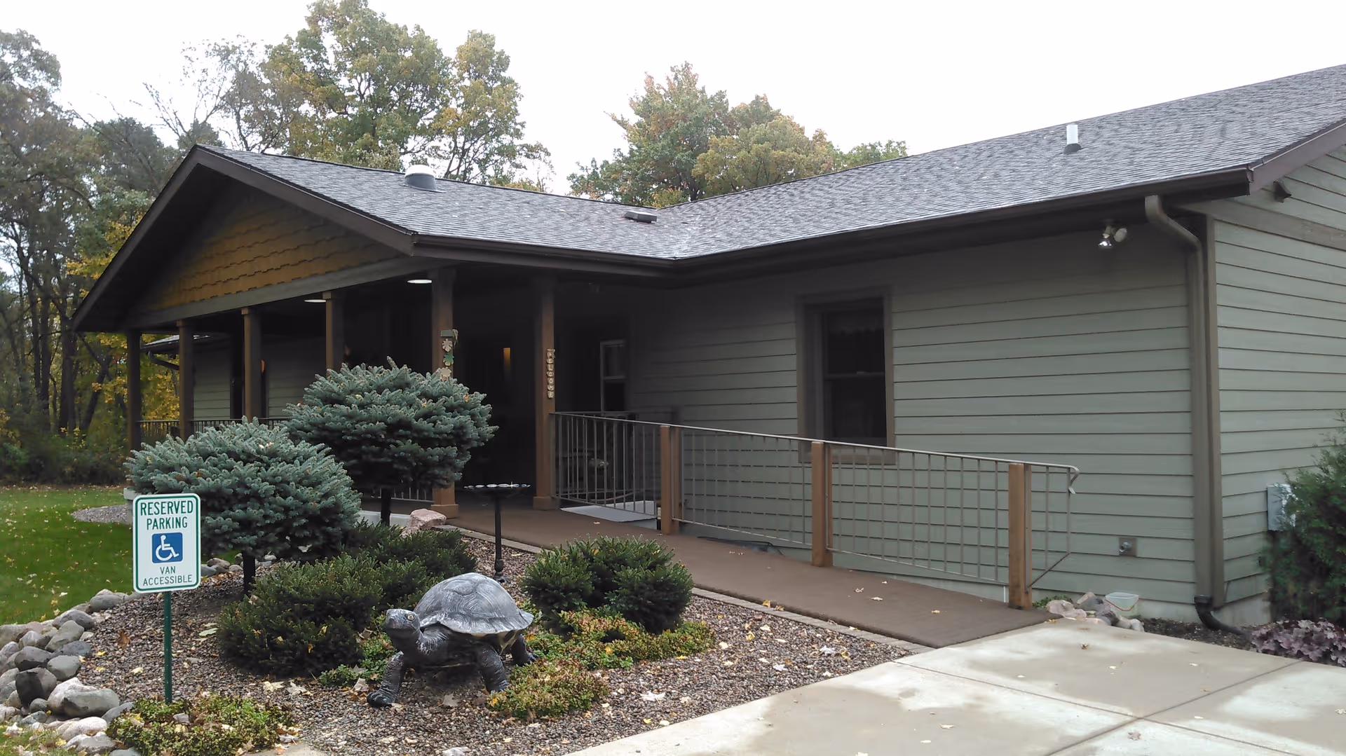 Exterior view of a single-story building with green siding and a gray roof, featuring a covered porch with wooden posts and a wheelchair accessible ramp. In front of the building, there is a landscaped area with bushes, a small tree, and a decorative turtle statue. A reserved parking sign for van accessible handicapped parking is visible near the landscaped area.