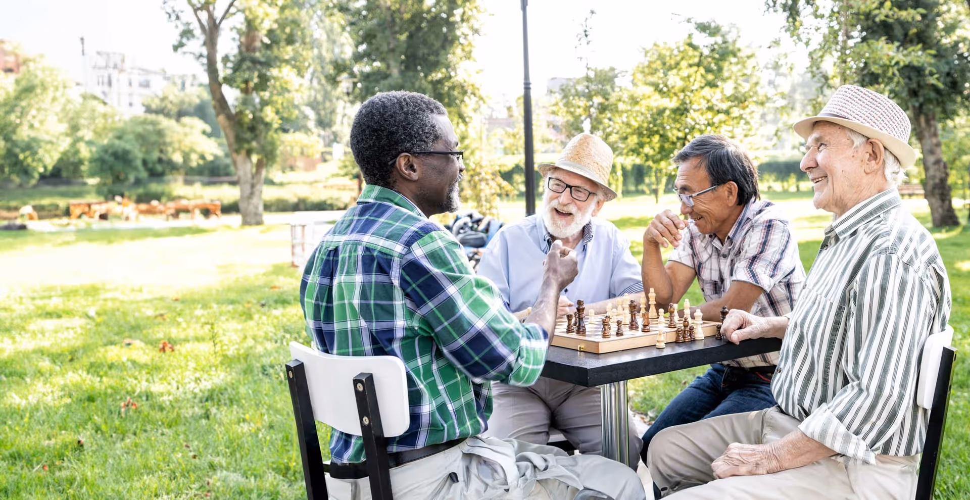 Four elderly men sit at a table outdoors playing chess in a park.