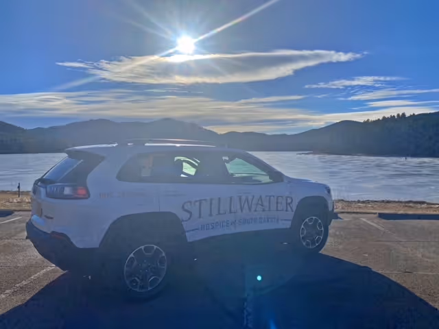 White SUV with Stillwater Hospice of South Dakota logo parked by a lakeside with mountains.