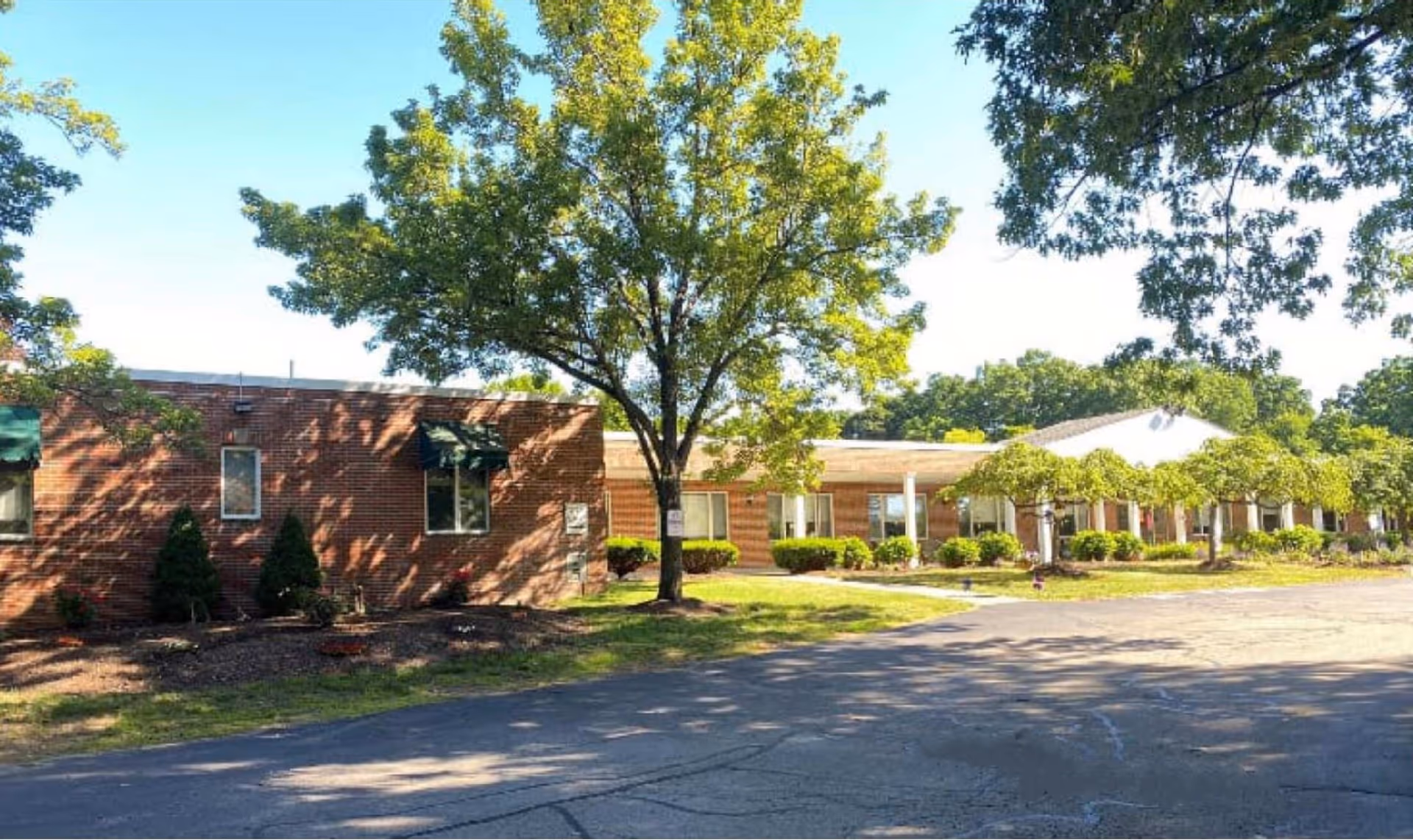 Exterior view of Charlwell House Health & Rehabilitation Center showing a single-story brick building with several windows and green awnings. There are trees and shrubs in front of the building and a paved driveway or parking area in the foreground under a clear blue sky.