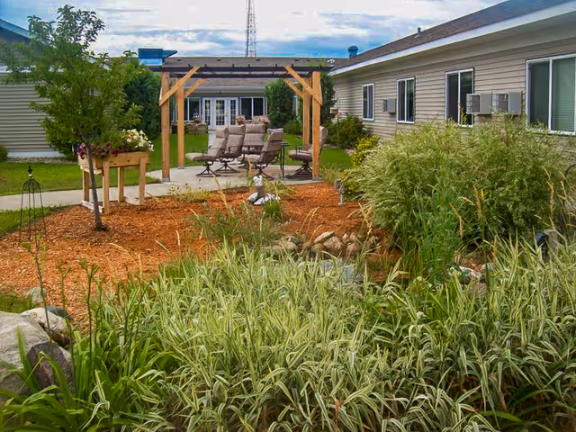 Outdoor garden area at Valley View Estates Assisted Living featuring a wooden pergola with patio chairs and a table, surrounded by various plants, shrubs, and a small water feature. The building with multiple windows is visible in the background under a partly cloudy sky.