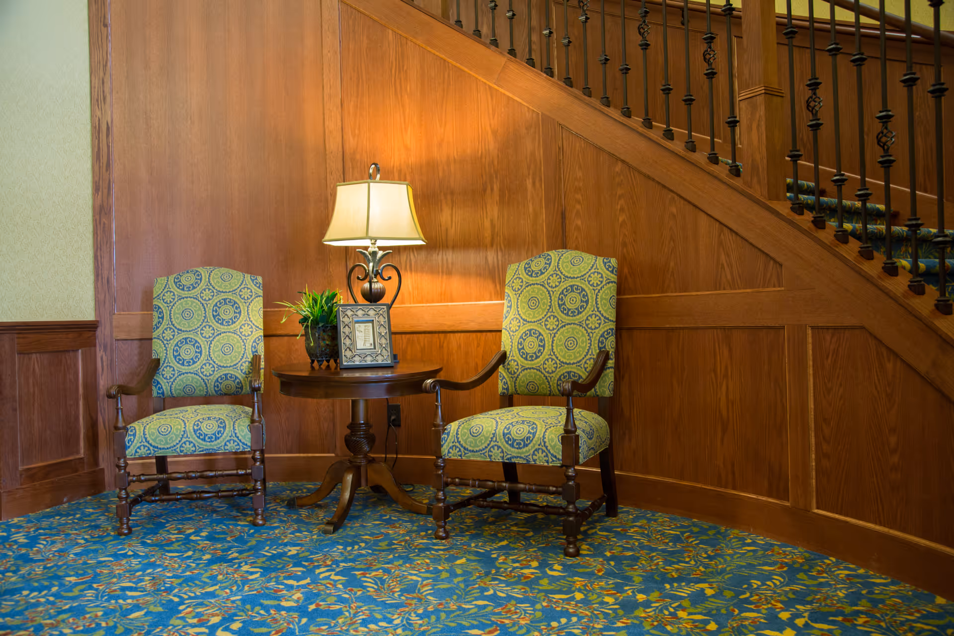Two upholstered armchairs flank a round table with a lamp beside a wooden staircase.