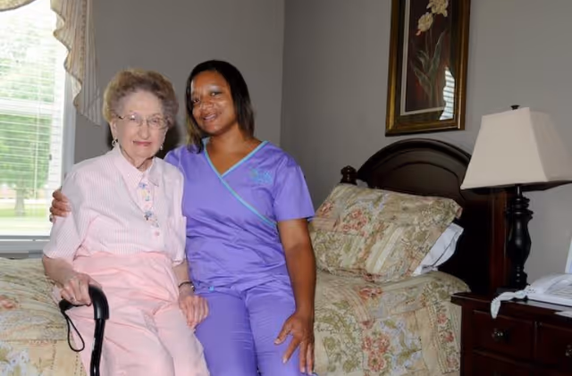 An elderly woman with glasses and a cane sitting on a bed next to a smiling caregiver in purple scrubs in a bedroom with floral bedding, a bedside lamp, and a framed flower picture on the wall.