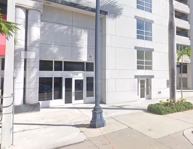 Modern beige building exterior with glass entrance doors and palm trees along the sidewalk.
