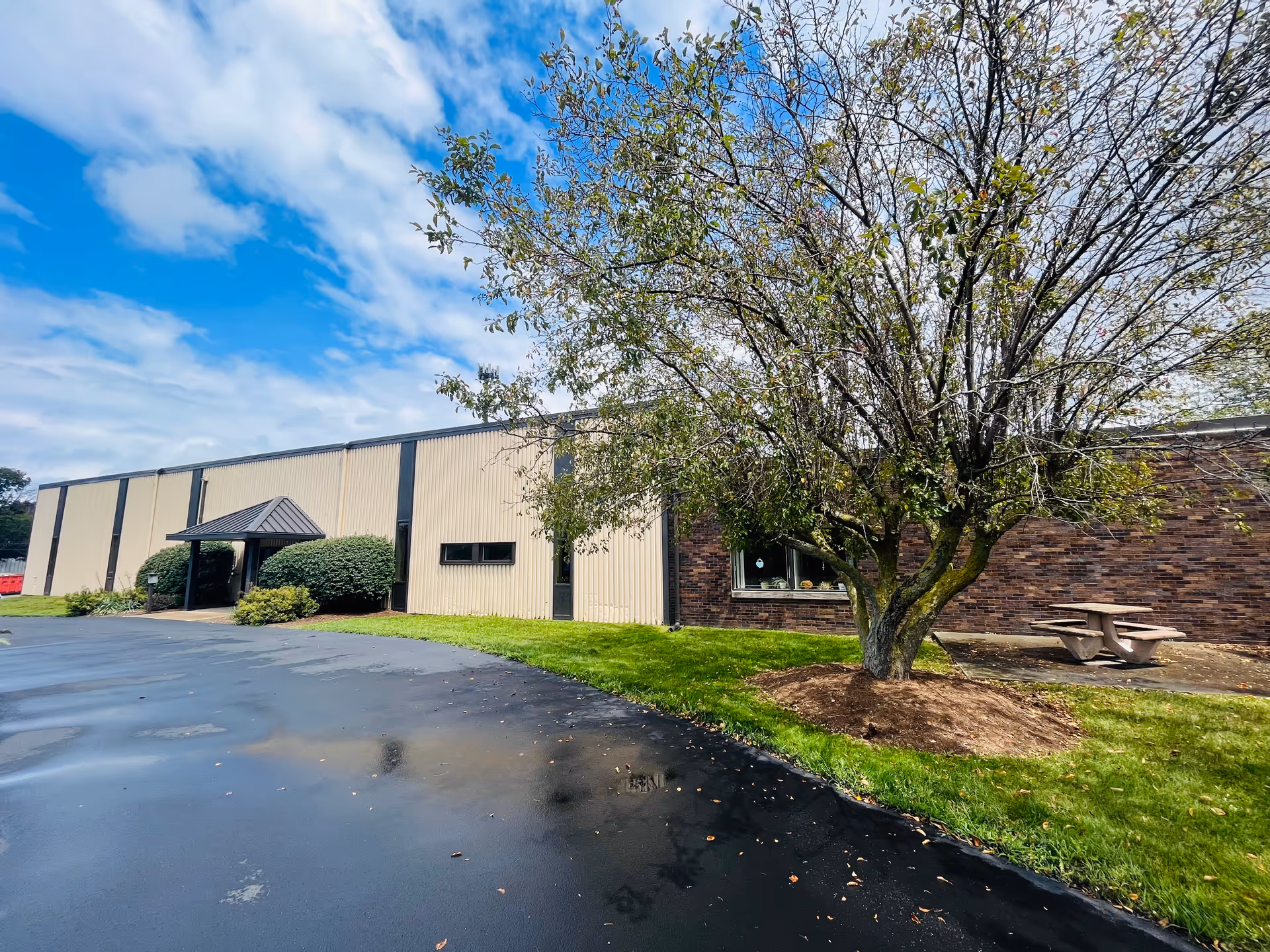Exterior view of a senior facility building with a tree, parking area, and picnic table.