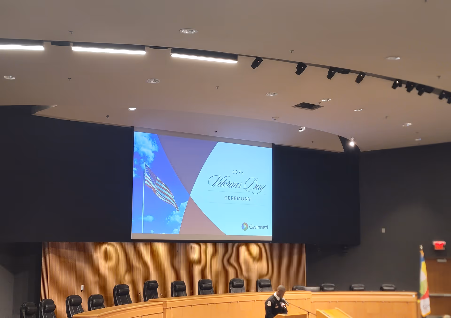 A large interior conference room features a projection screen displaying a Veterans Day ceremony.