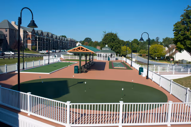 Outdoor recreational area at Phoebe Berks Health Care Center featuring a fenced putting green, shuffleboard courts, a covered seating area with benches, street lamps, and a white gazebo in the background surrounded by trees and nearby buildings.