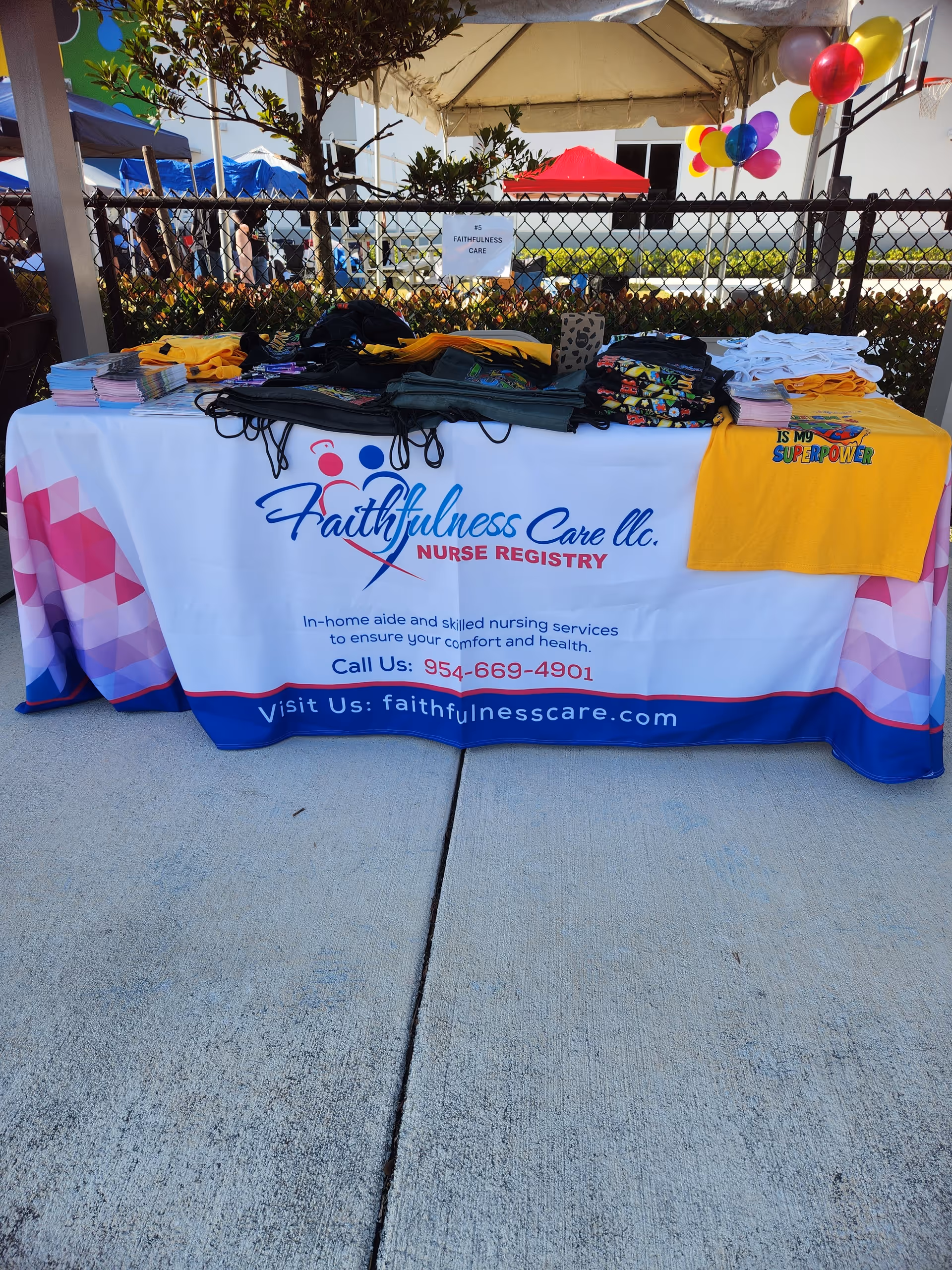 Outdoor table with Faithfulness Care LLC banner, shirts, and balloons at an event.