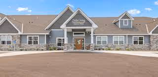Front exterior of a single-story senior living facility with gray siding, stone accents, a circular driveway, and a centered covered entrance with a sign reading 'Suite Living Lakeville'.