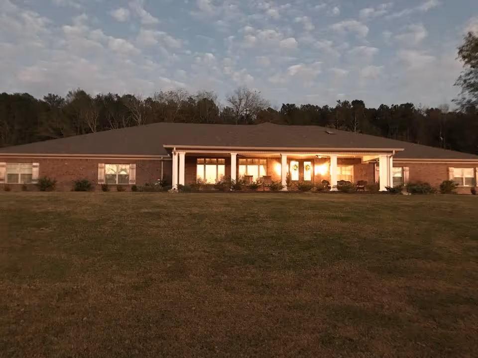 Exterior front view of a single-story brick building with a large covered porch and several windows, surrounded by a grassy lawn and trees in the background under a cloudy sky.