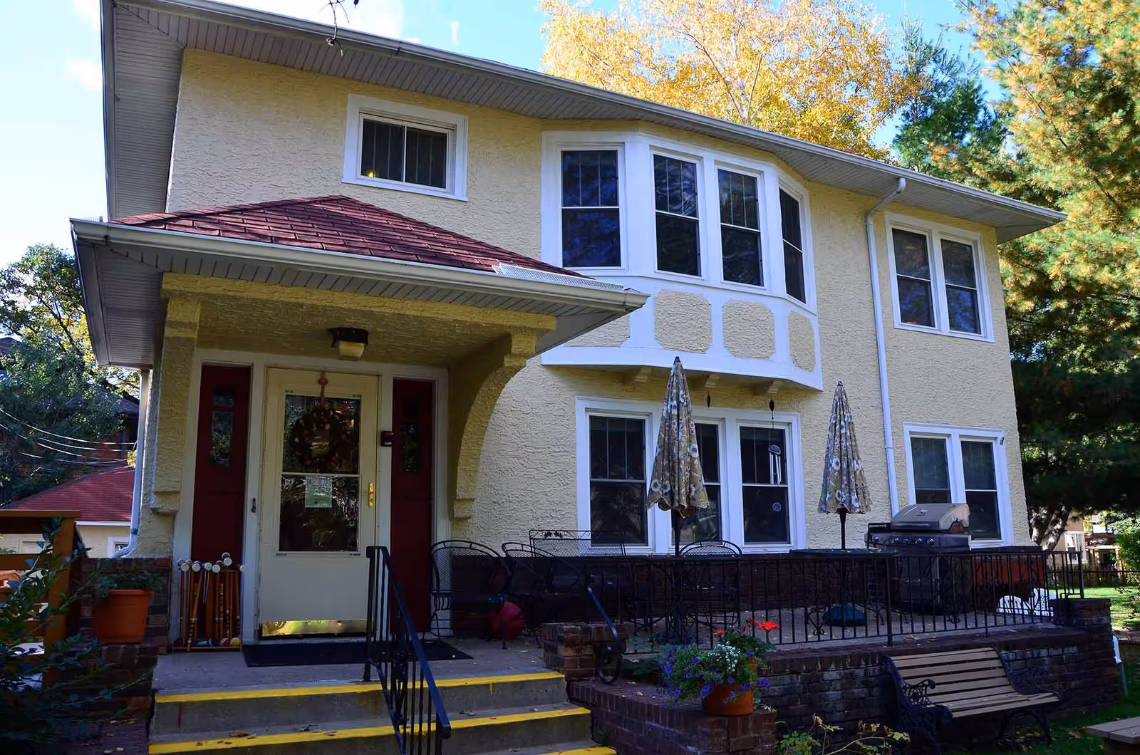 Exterior view of a two-story residential building with a yellow textured facade and white trim around the windows. The building has a small porch with steps leading up to a white door with glass panels. There are two patio umbrellas and outdoor furniture on a raised brick patio area to the right of the entrance. Trees and greenery surround the building.