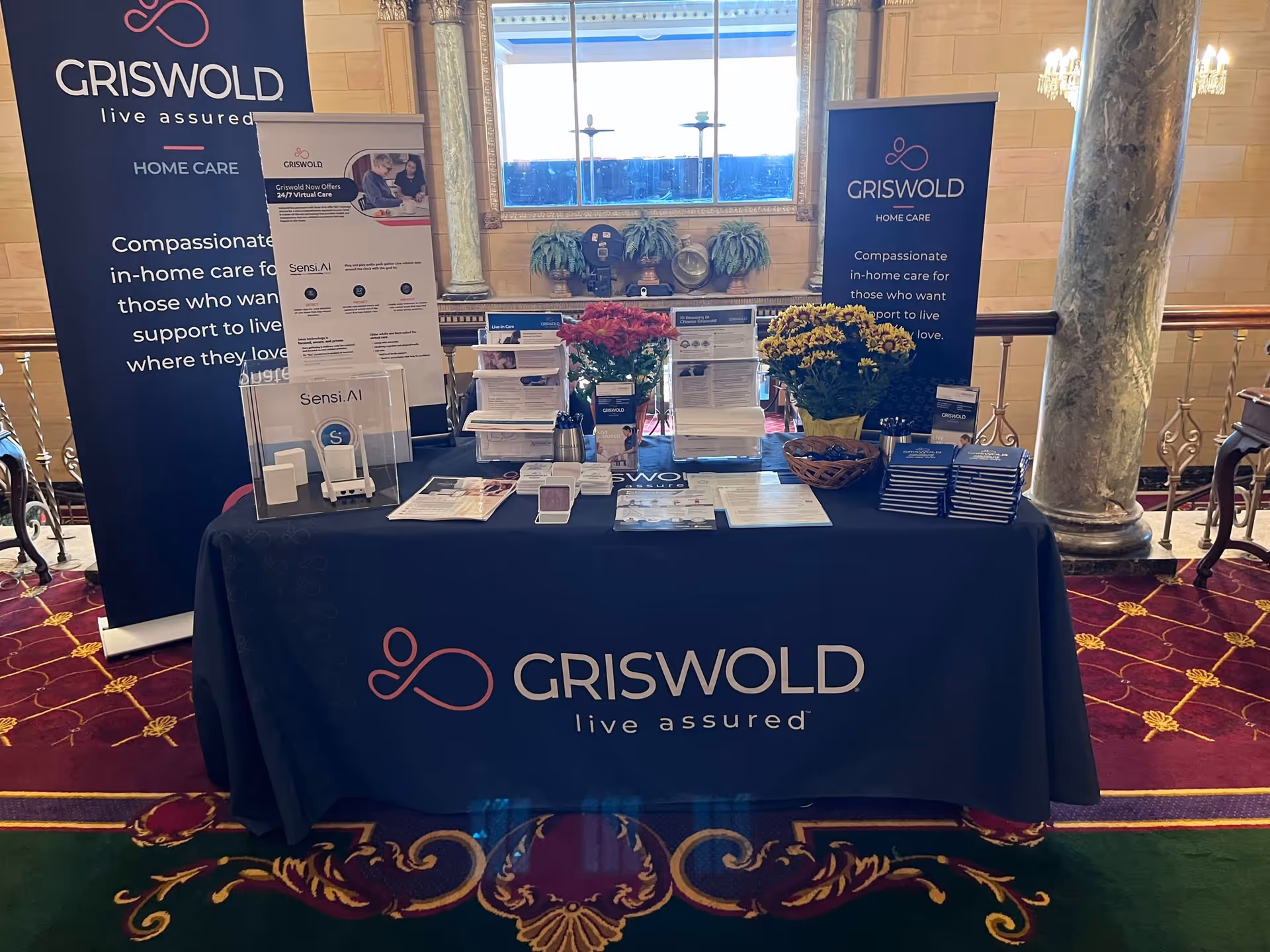 Booth setup with Griswold Home Care banners and a brochure-filled table in a lobby.