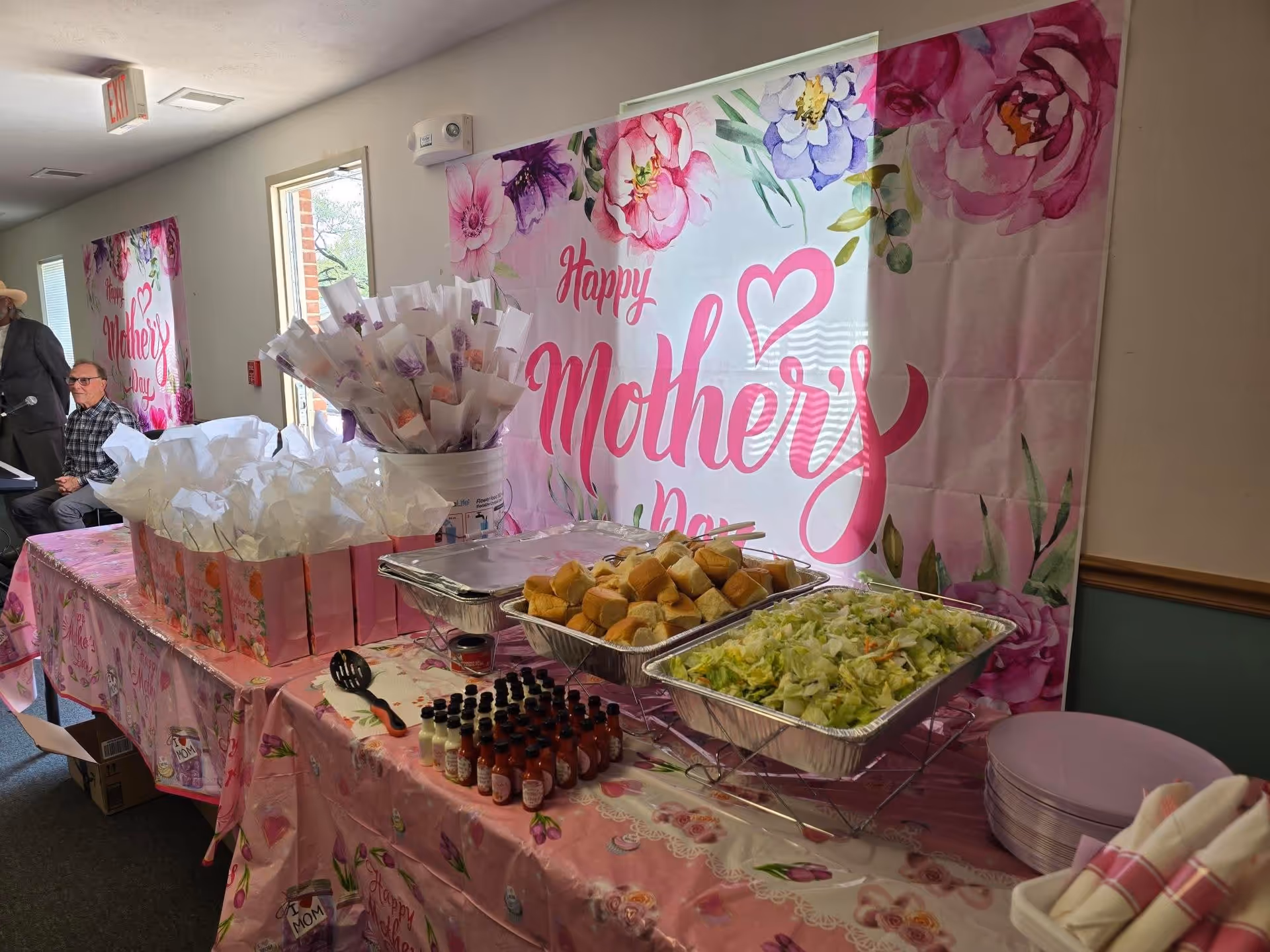 Indoor celebration table with pink Mother's Day backdrop, food trays, and guests visible in background.