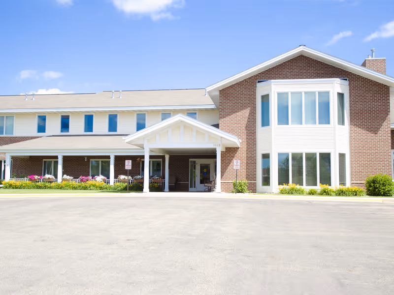 Front exterior view of a two-story senior living facility building with a covered entrance, large windows, and a brick and siding facade under a blue sky with some clouds.