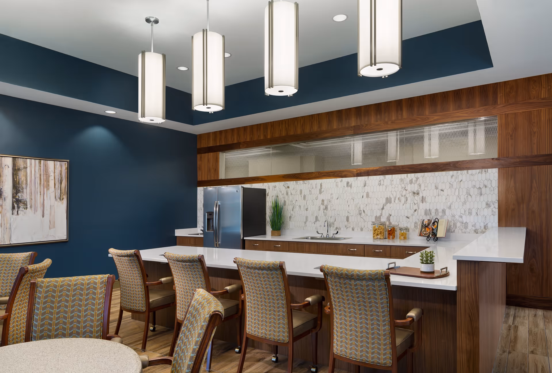 Modern communal kitchen and dining area with a long white countertop bar, upholstered chairs, pendant lights, and a stainless refrigerator.