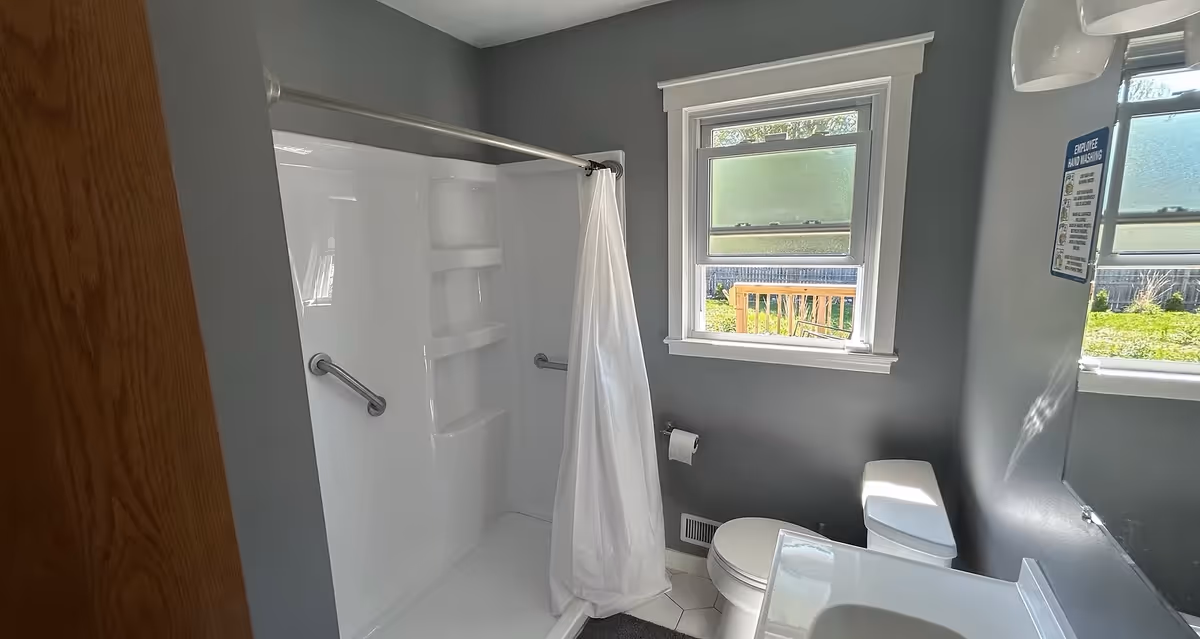 Small bathroom with a white shower stall, toilet, sink, grab bars, and two windows.