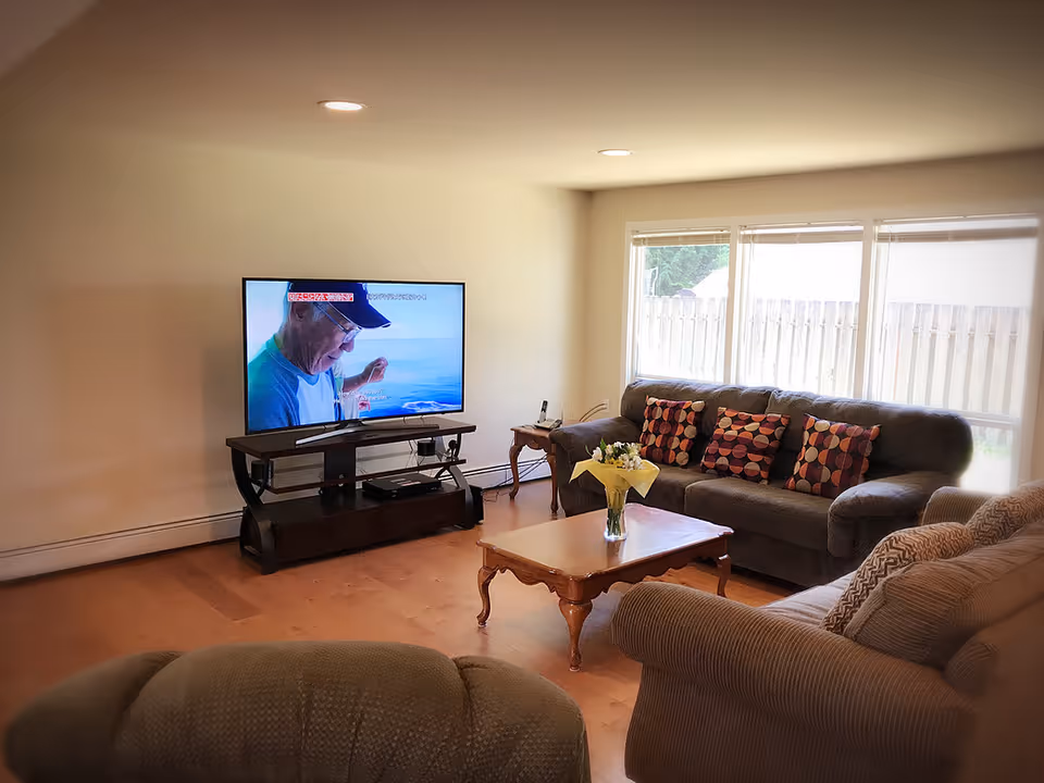 A cozy living room with a large flat-screen TV on a dark wooden stand, a wooden coffee table with a vase of flowers, a dark gray sofa with patterned cushions, and a beige armchair. Large windows with blinds allow natural light to fill the room.