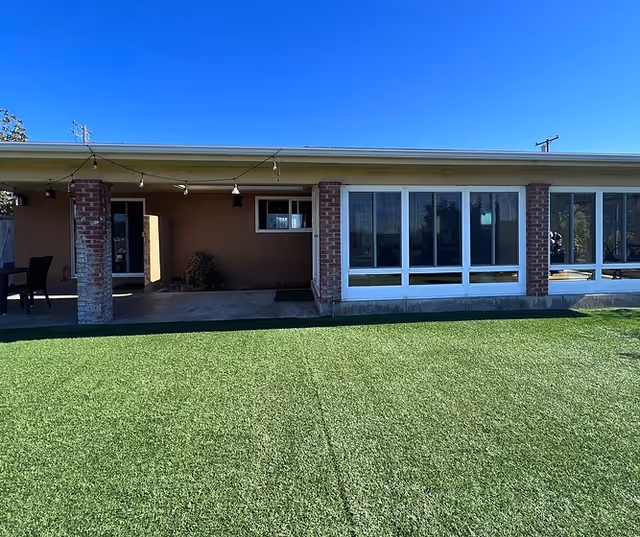 Single-story brick and stucco building with large windows and a covered patio overlooking a green artificial turf lawn under a clear blue sky.