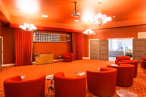 A spacious room with bright red walls, ceiling, and carpet featuring a patterned design. The room contains several red upholstered chairs arranged in a semi-circle facing a wooden cabinet and a large bingo board mounted on the wall. There are three chandeliers hanging from the ceiling providing warm lighting. Double doors open to an adjacent room with neutral-colored furniture.