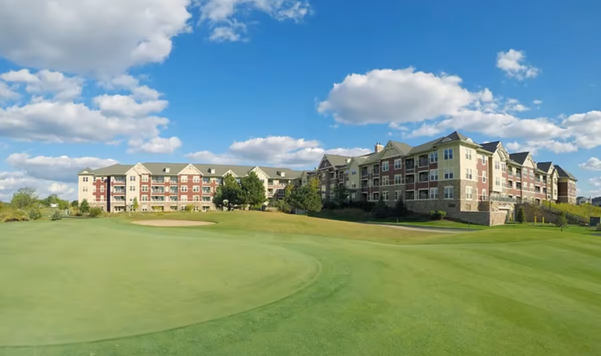 Wide exterior view of a large senior living facility building with multiple floors, set against a blue sky with scattered clouds. The building is surrounded by well-maintained green lawns and landscaping, resembling a golf course or open grassy area in the foreground.