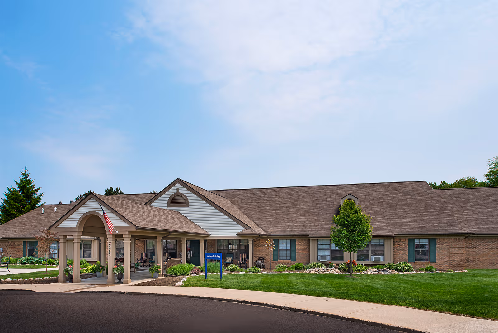 Exterior view of a single-story senior living facility building with a covered entrance, an American flag, well-maintained lawn, small trees, and a clear blue sky.