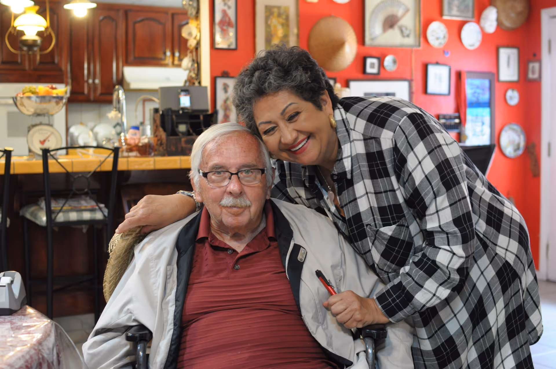 An elderly man sitting in a wheelchair with a woman standing beside him, smiling and leaning in for a hug. They are indoors in a warmly decorated room with red walls adorned with various framed pictures and decorative plates. A kitchen area with wooden cabinets and a counter is visible in the background.