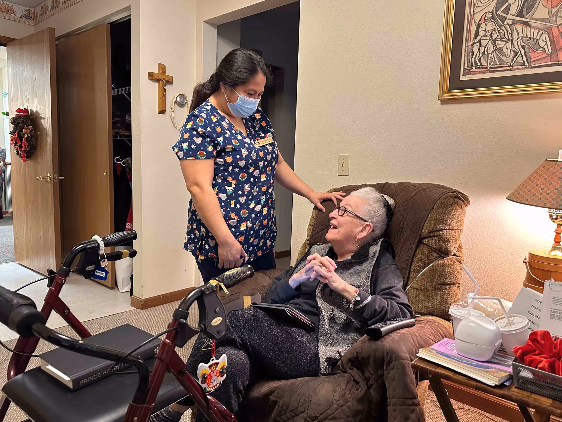 A caregiver wearing a festive scrub top and a face mask is standing next to an elderly woman seated in a recliner chair. The elderly woman is smiling and looking up at the caregiver. There is a walker with a book on it in the foreground, a side table with a lamp, cups, and books, and a framed artwork on the wall behind them. The setting appears to be a cozy living room or common area in an assisted living facility.