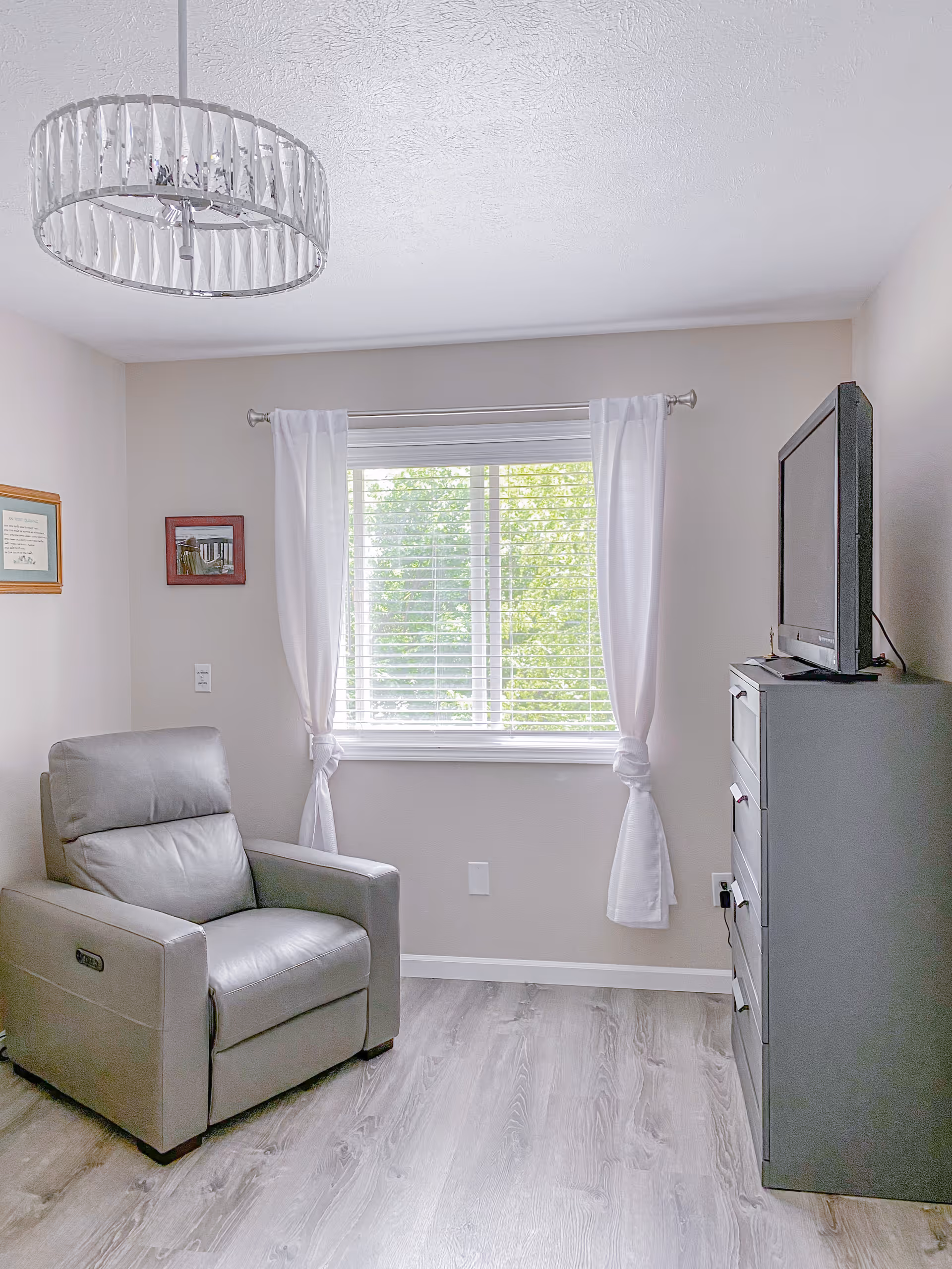 Small bright living area with a gray recliner, window, curtains, and dresser with TV.