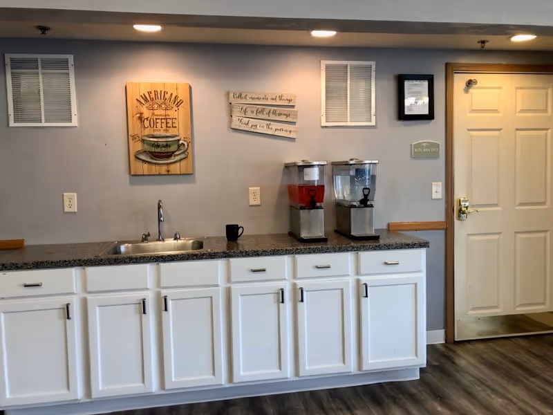 Kitchen area with white cabinets, countertop, sink, two beverage dispensers, and wall art.